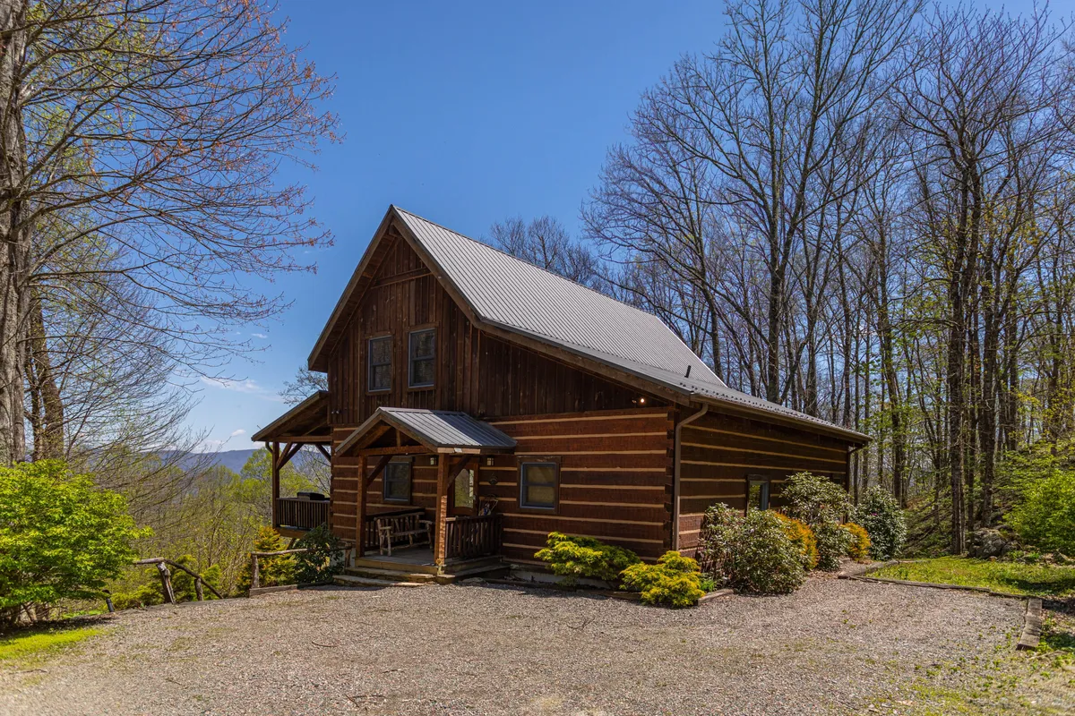 This image showcases a charming log cabin nestled amidst lush greenery, with a spacious front porch perfectly positioned for taking in sweeping mountain sunsets. The peaceful setting is inviting, with comfortable seating on the porch and plenty of space for relaxation or gathering with loved ones.