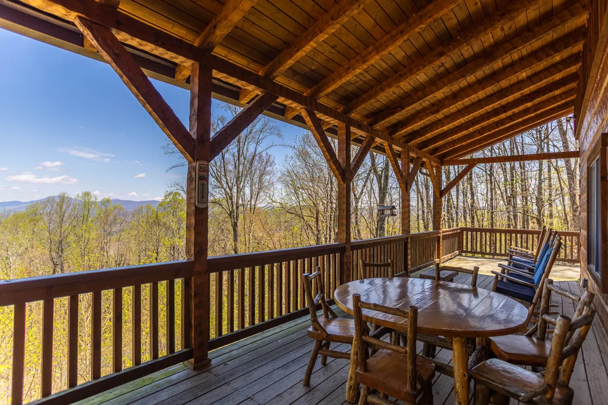 An expansive covered wooden deck featuring a round table with rocking chairs, all oriented to maximize views of the surrounding mountains and landscape. The scene exudes tranquility and encourages enjoying coffee at sunrise or sunsets with friends and family.