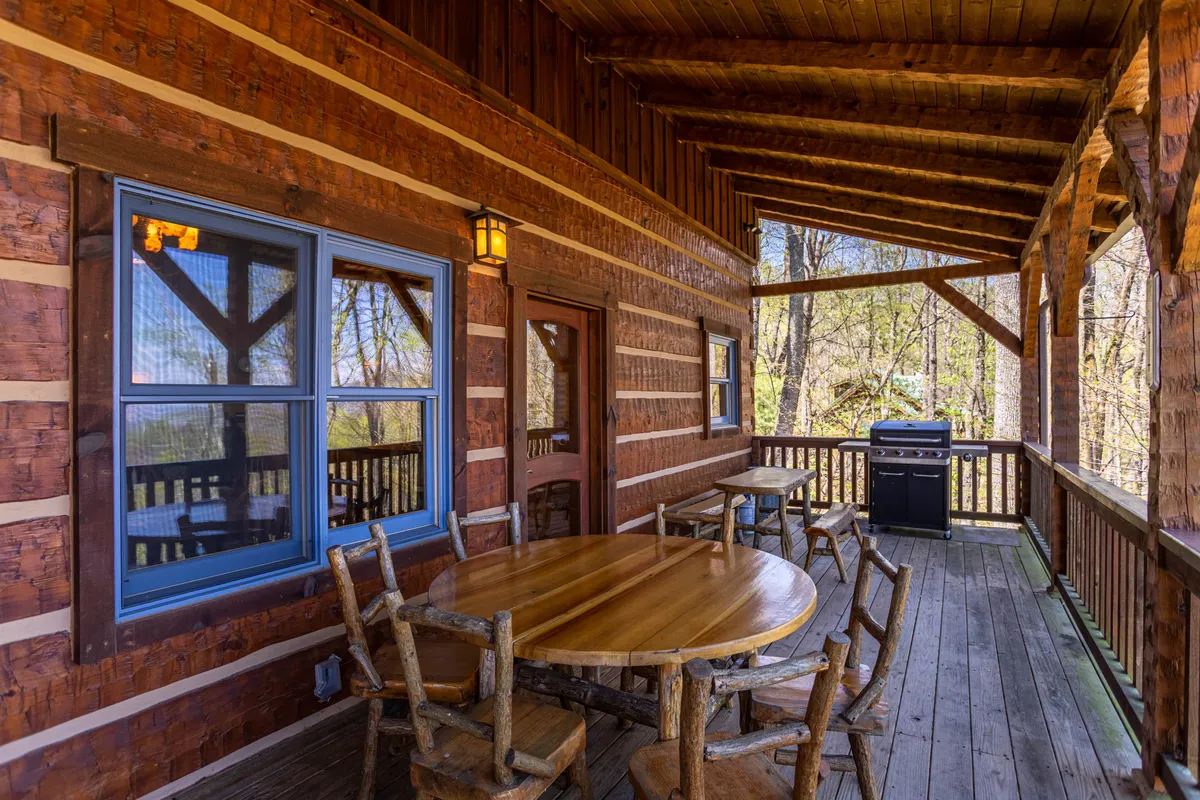 The image shows the exterior of the cabin, featuring a wooden table and grill set on a porch, surrounded by lush green trees. The log cabin structure and natural setting invite viewers to imagine al fresco meals and gatherings in a tranquil mountain landscape.