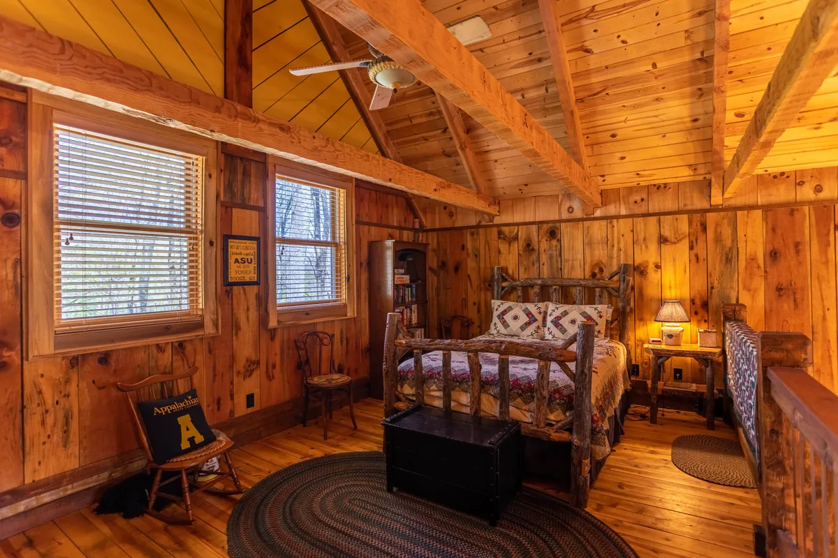 A rustic bedroom featuring a handmade chair next to a window, a bookshelf, and a table lamp. The scene is set for quiet reflection or birdwatching, with natural light streaming in and a clear view outside.