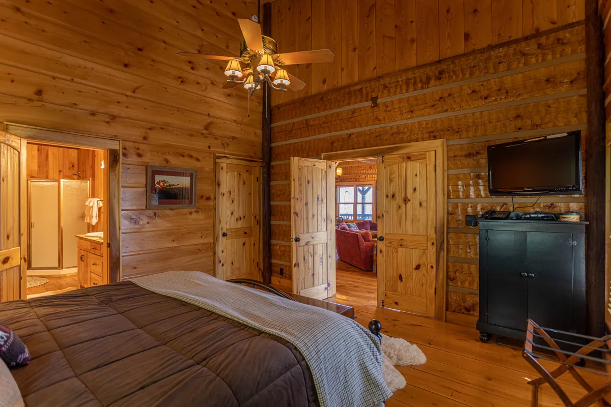 A cozy bedroom with wooden walls, a bed layered with soft blankets, doorways leading to other rooms, a ceiling fan, and a television. The space feels comfortable and inviting, ideal for relaxation after a day of adventure.