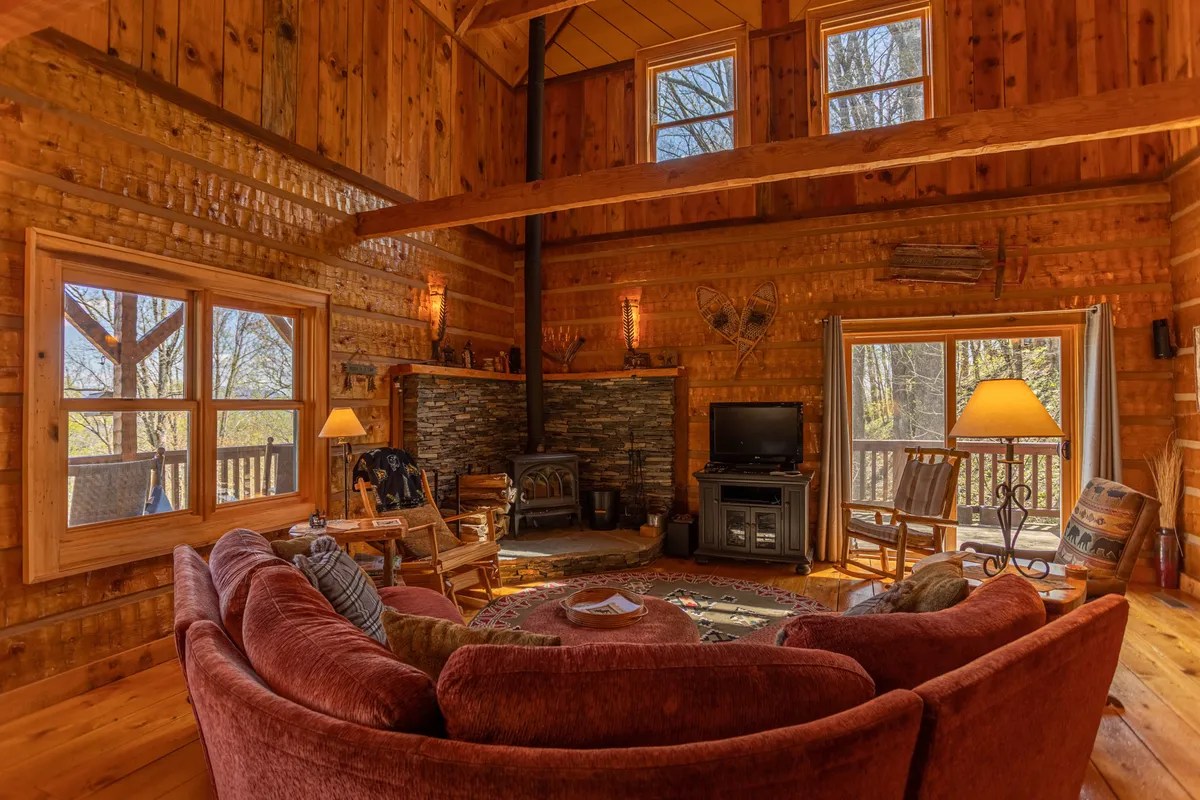 A spacious living area featuring a large round sofa centered around a striking stone fireplace, with exposed wooden beams overhead and expansive windows framing the mountain landscape. The ambiance is cozy, inviting guests to relax, converse, or enjoy the warmth of the fire while surrounded by nature-inspired design elements.