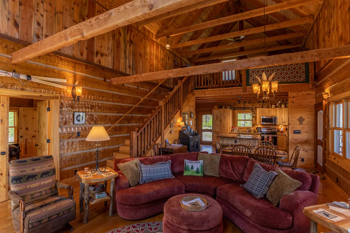 A spacious, open-plan living area with a red sectional sofa, a prominent wooden staircase, rustic lighting, and large windows that pull in the winter scenery. The scene emphasizes gathering, relaxation, and the natural connection to the outdoors.