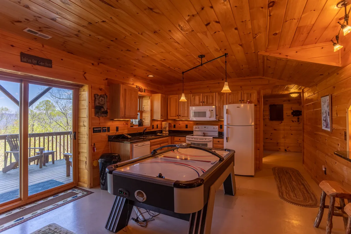This image showcases a lively recreation area featuring an air hockey table, kitchen appliances visible in the background, wooden ceilings, and large windows. The setup suggests an open concept space designed for play, casual dining, and socializing, all flooded with natural light from the deck access.