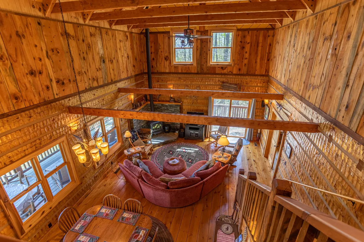 A spacious living area featuring prominent wooden beams, a round pink sofa, large windows, and a dining table. The expansive windows frame views of the surrounding trees and let in abundant natural light, creating an airy atmosphere that invites guests to experience the mountain environment from within.