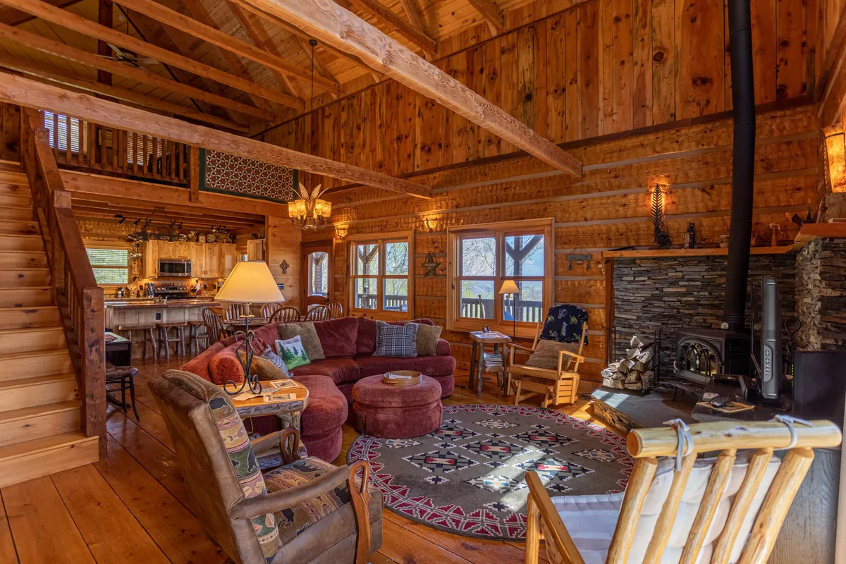 A warm, inviting great room featuring a large circular couch positioned near a striking stone fireplace, with handcrafted rugs and a wooden staircase visible. The background reveals a dining area and glimpses of the kitchen, all beneath exposed wooden beams and large windows that let in natural light and frame the mountain views.