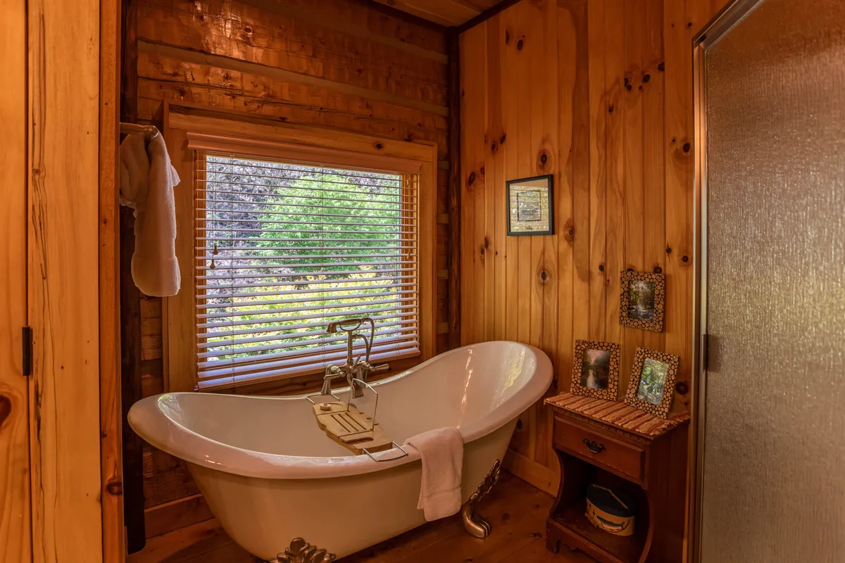 A serene bathroom showcasing a freestanding claw foot bathtub, a window with blinds for privacy, wood-paneled walls, and decorative pictures. The peaceful ambiance invites long, soothing soaks and moments of solitude.