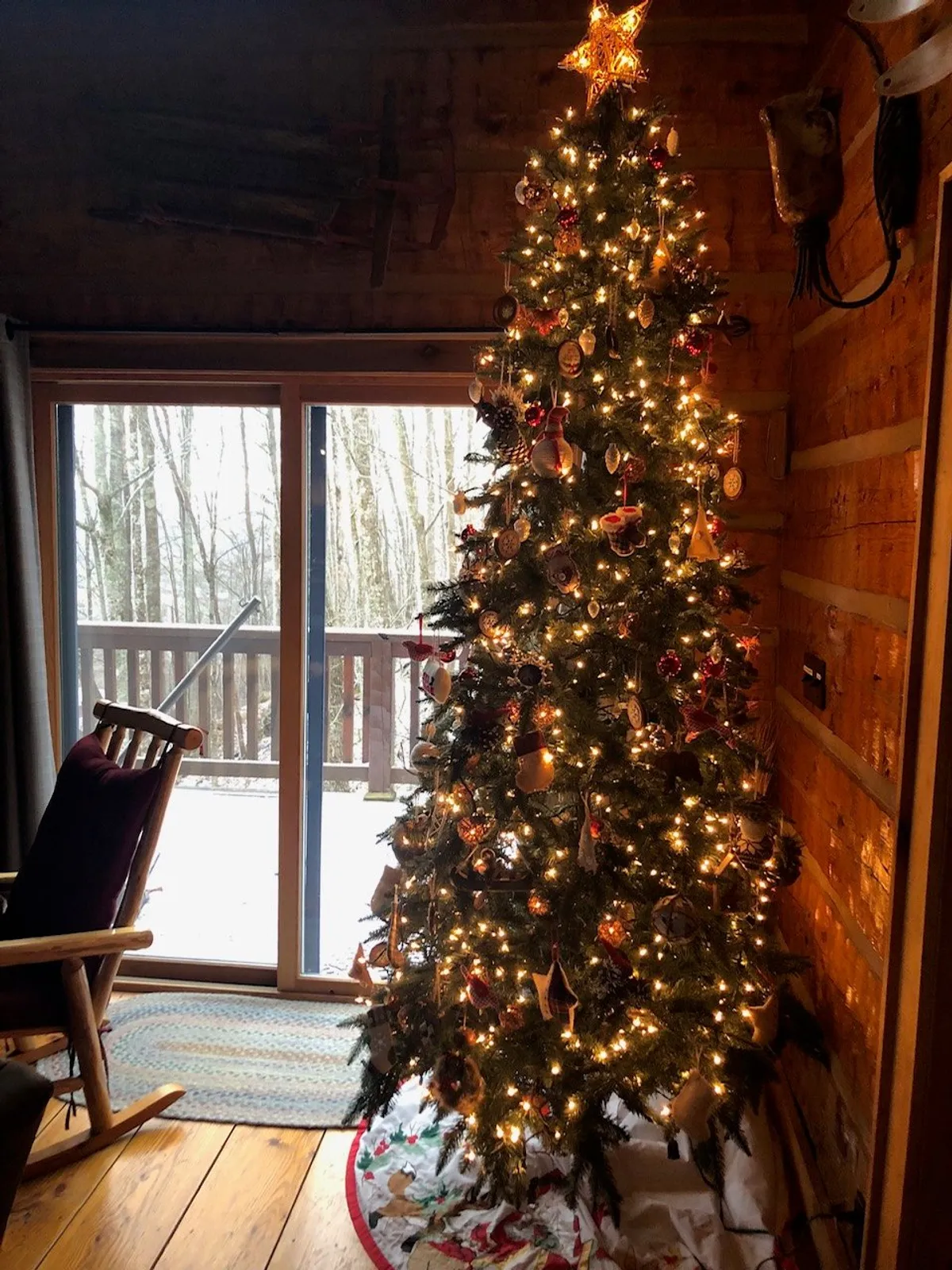 This image captures a warmly decorated living area with a Christmas tree, a rocking chair positioned by a large window, and inviting wintertime scenery outside. The window offers a glimpse of snow-covered grounds, linking the cozy interior directly to the natural landscape outside.